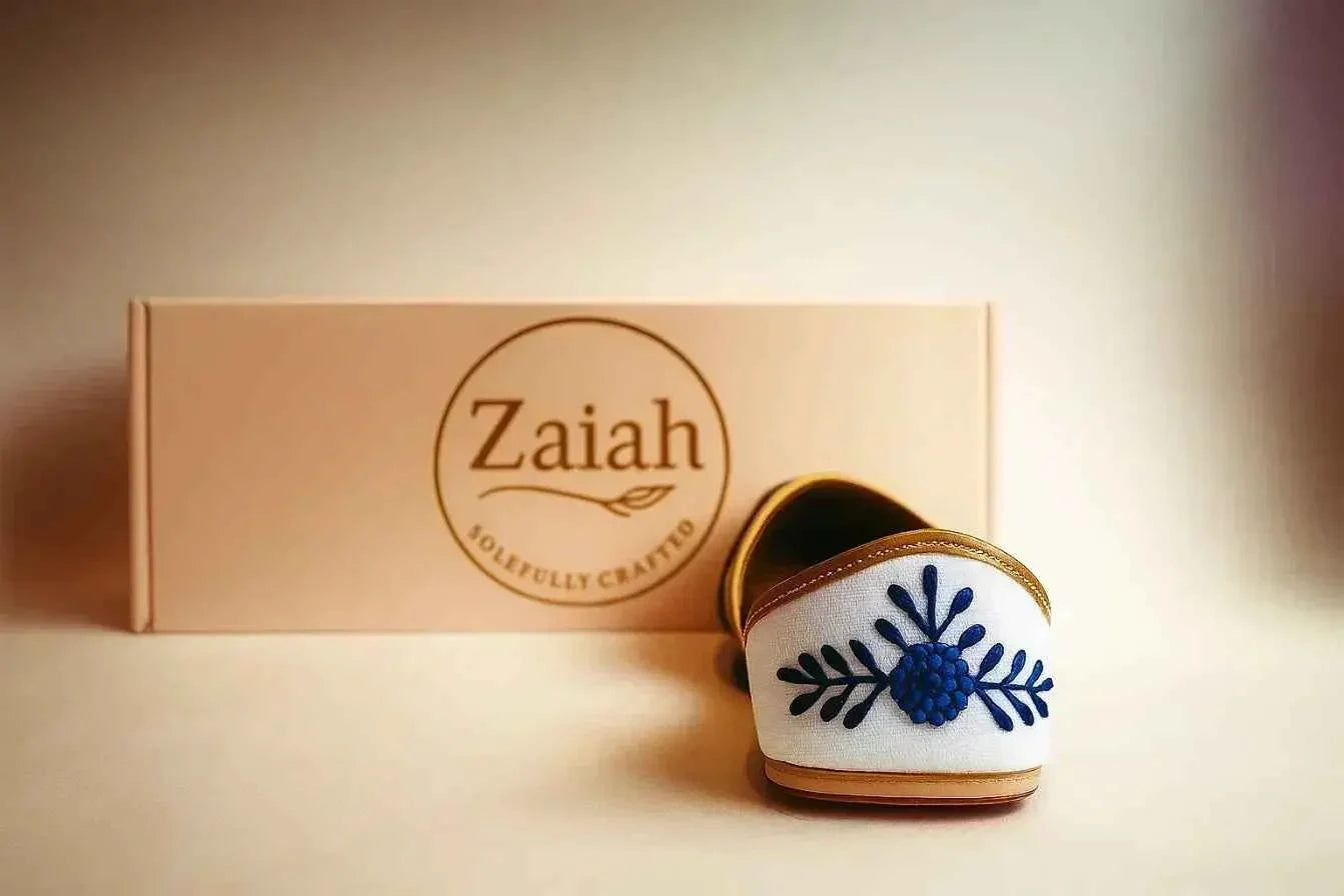 White handcrafted jutti with blue floral embroidery, elegant slip-on design, displayed with branded box.