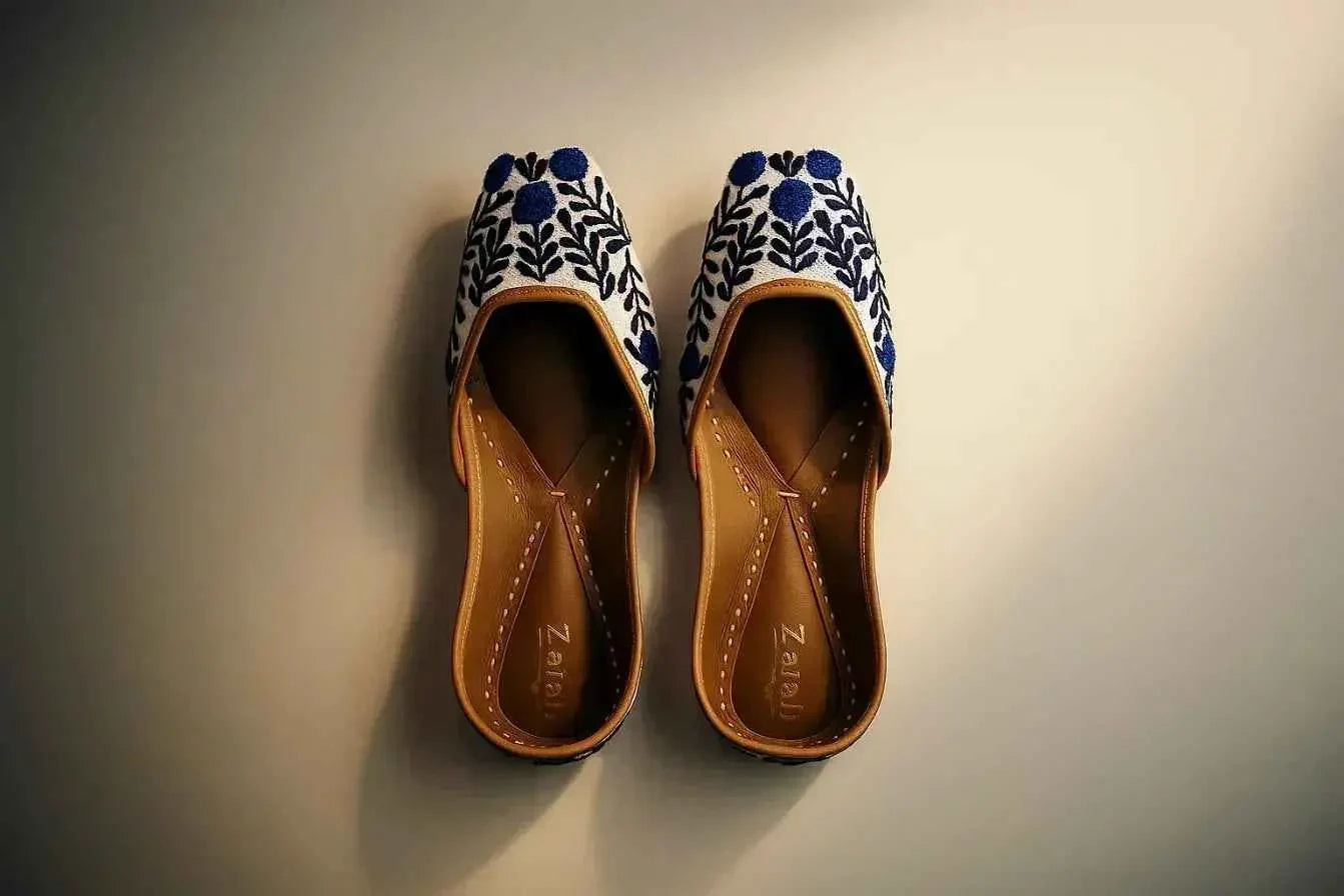 Handcrafted white juttis with blue floral embroidery, tan leather soles, elegant traditional footwear