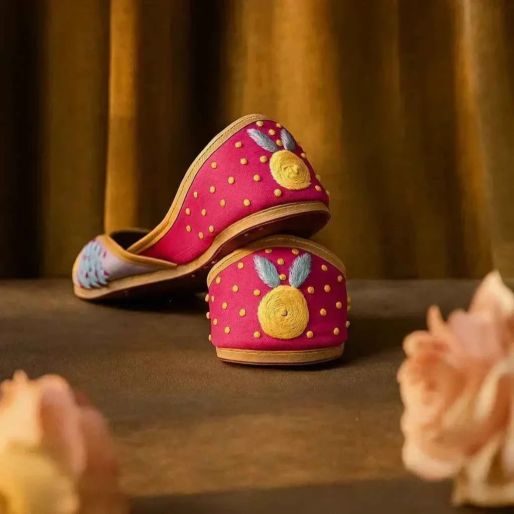 Handcrafted pink juttis with yellow floral embroidery and gold dots, styled for romantic occasions.