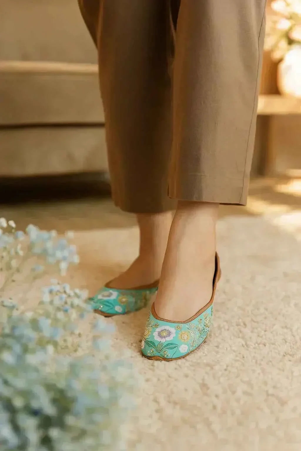 Turquoise floral embroidered juttis with gold detailing, styled with cropped trousers on a soft rug