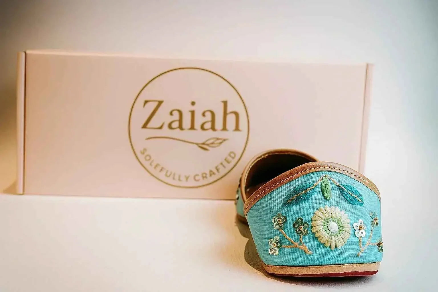 Turquoise handcrafted jutti with floral embroidery, displayed beside Zaiah branded box