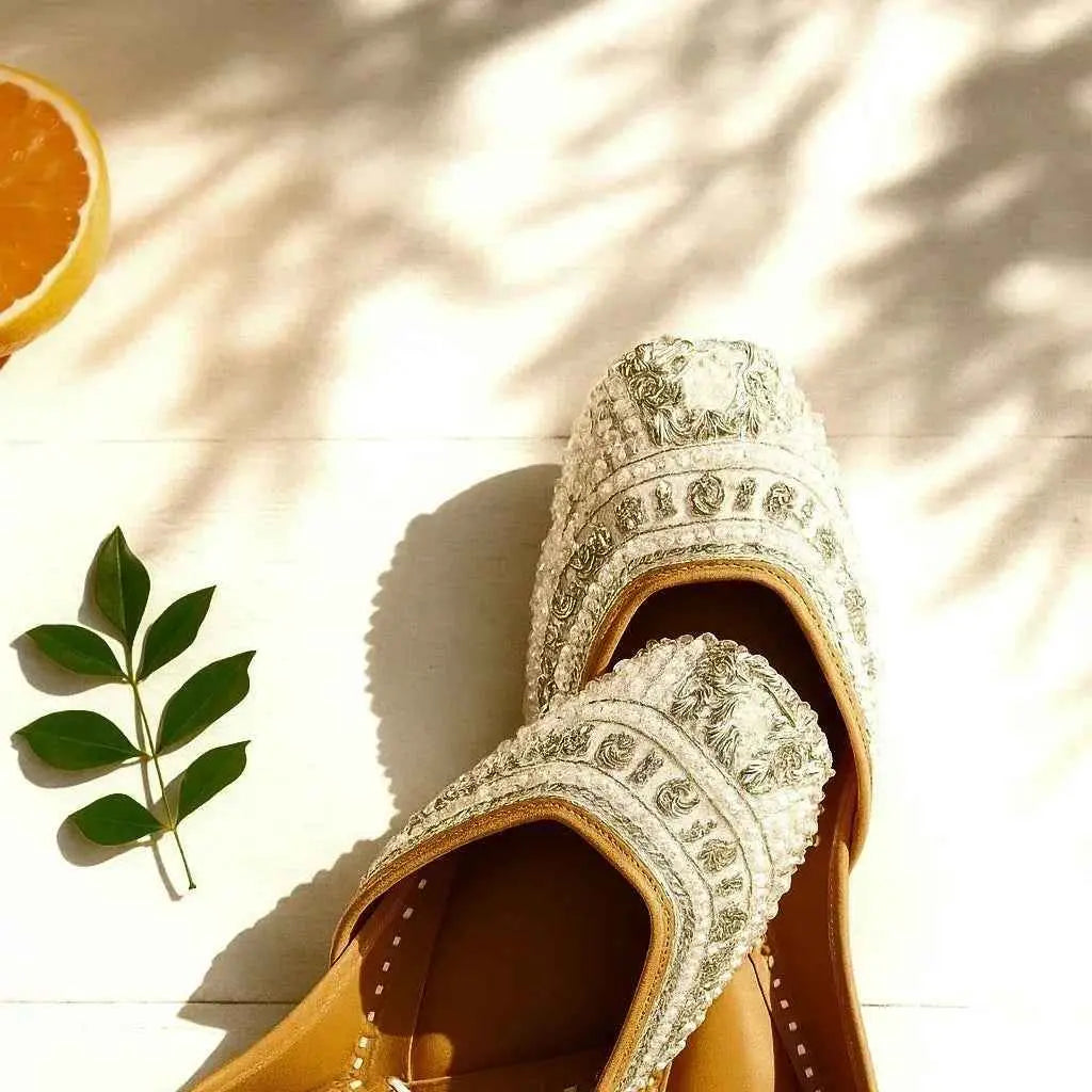 Handcrafted embroidered juttis with floral detailing, styled with orange slice and green leaf under sunlight